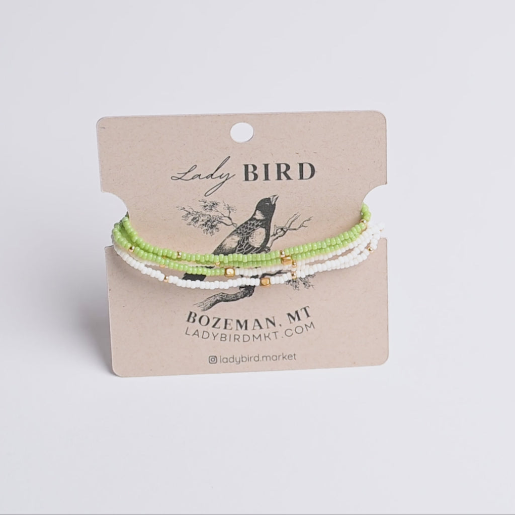 Three beaded bracelets in light green, white, and gold tones displayed on a brown kraft card labeled Lady Bird, photographed against a light neutral background.