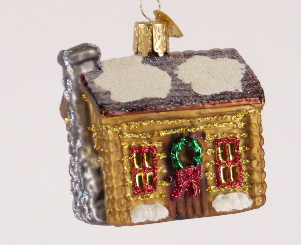 Glass ornament of a cabin with snow on the roof and Christmas decorations on a white background