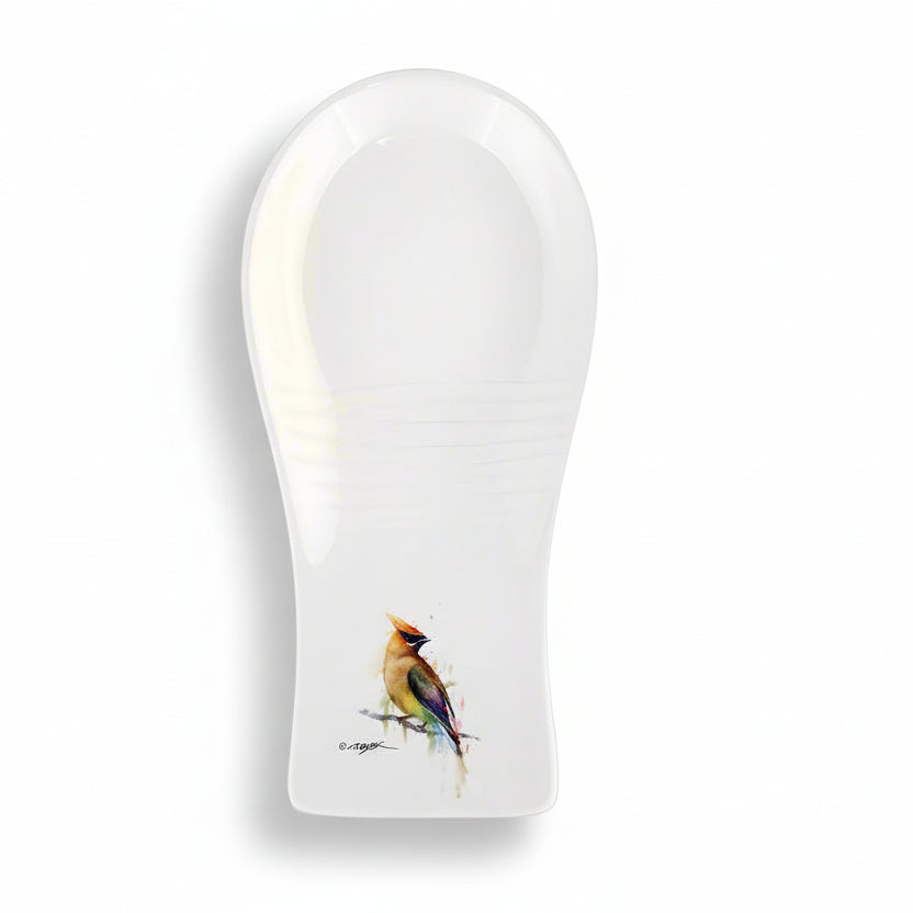 A white stoneware spoon rest with a colorful bird, the Cedar Waxwing, perched on the side.